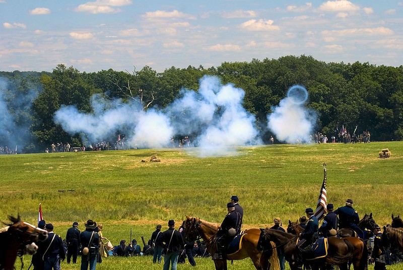 Field with Union soldiers on horses in foreground, smoke ad opposing soldiers along a tree-line in background