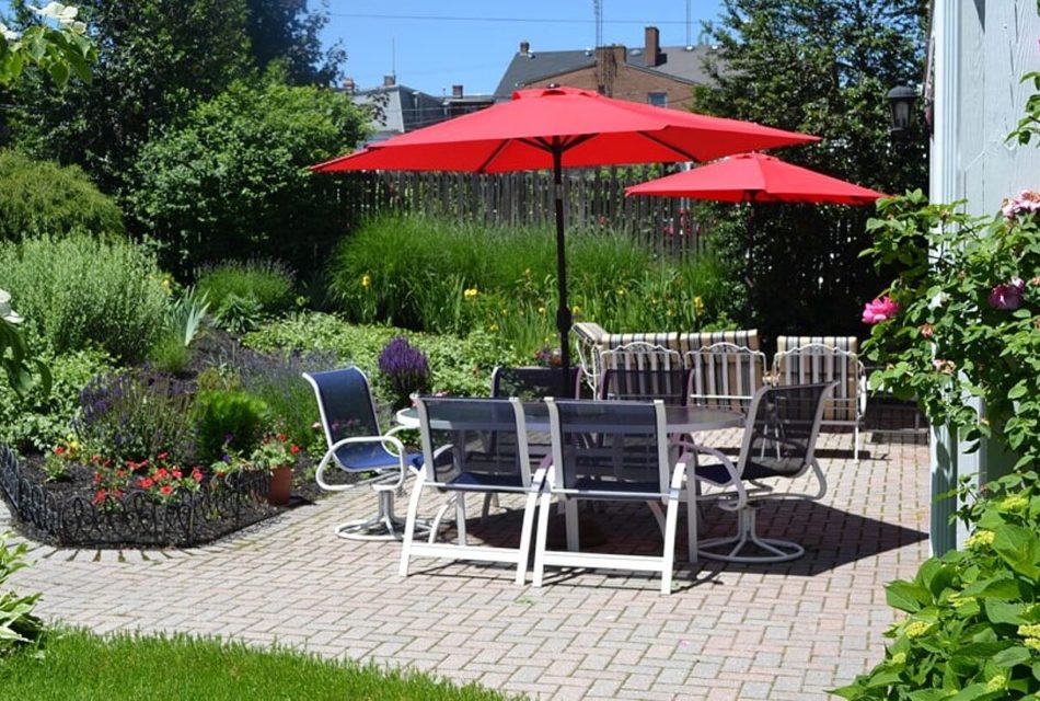 Backyard brick patio with lush landscaping and two patio tables with chairs and red umbrellas