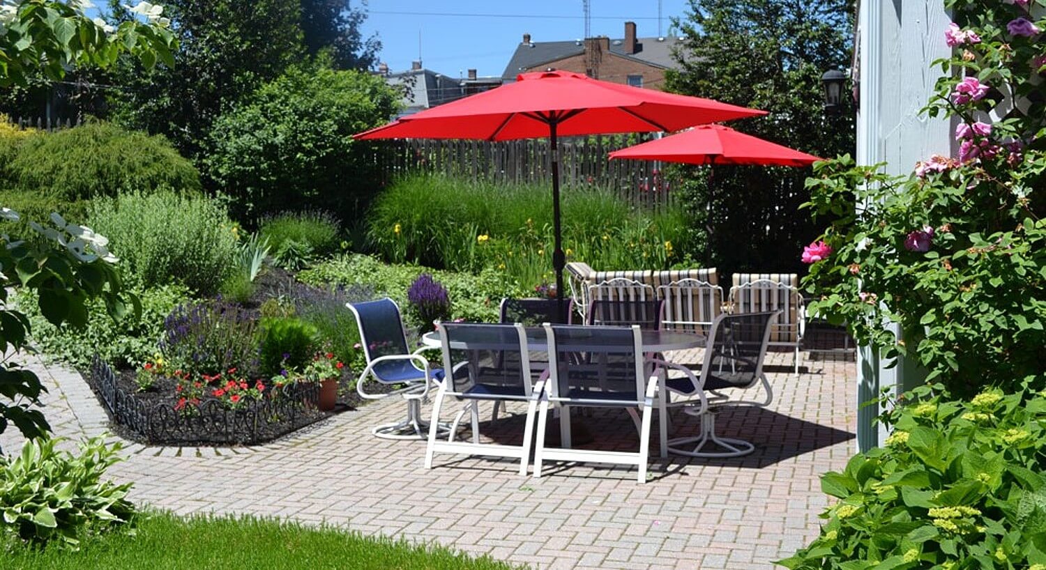 Backyard brick patio with lush landscaping and two patio tables with chairs and red umbrellas