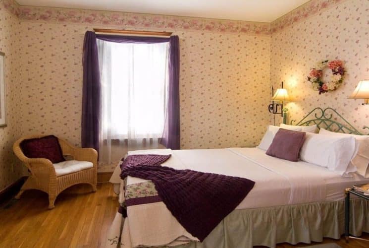 Cozy guest room with queen bed, flowered wallpaper, large window and wicker sitting chair