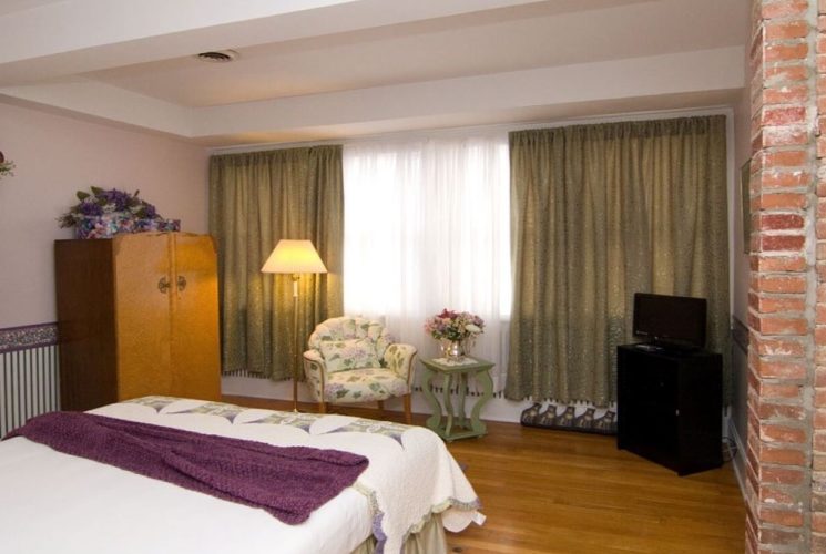 Large bedroom with queen bed, armoire, chair, and tall lamp in front of a window