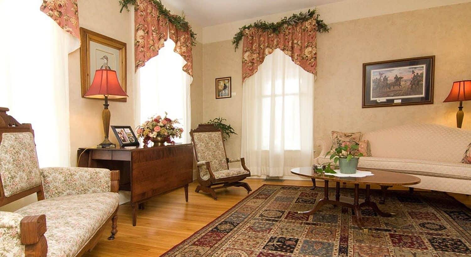 Living room of a home with a cream colored couch, two chairs, coffee table and three windows with floral curtains