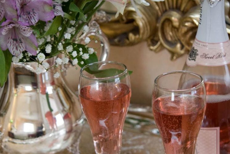 Two champagne glasses with a bottle of rose on a silver tray with a teapot of flowers